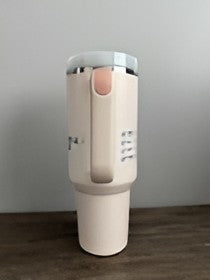 A tall, cylindrical insulated tumbler in a warm off-white or cream color, featuring a matching cream handle and a light gray lid, standing on a dark wooden surface against a plain gray wall.