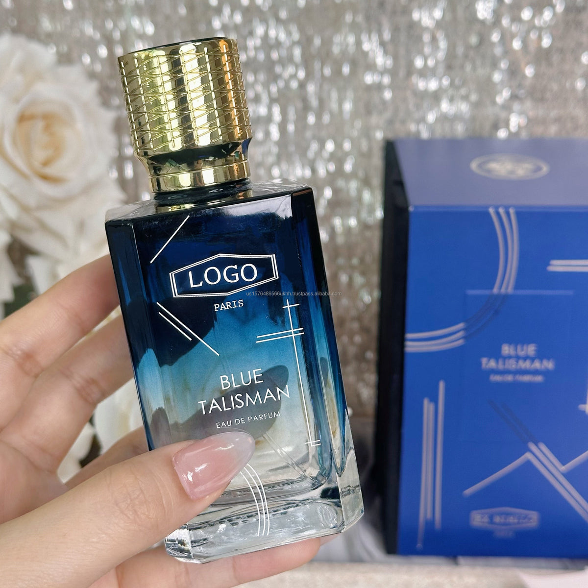 A hand holds a rectangular glass perfume bottle labeled 'BLUE TALISMAN' with a textured gold cap, next to its blue box packaging against a sparkly silver background.