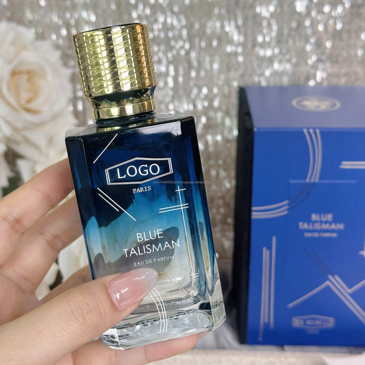 A hand holds a rectangular glass perfume bottle labeled 'BLUE TALISMAN' with a textured gold cap, next to its blue box packaging against a sparkly silver background.