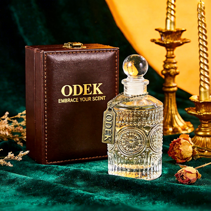 A clear glass perfume bottle with ornate cut-glass detailing and a round glass stopper, shown beside a brown leather ODEK box on a dark green velvet surface. Gold candlesticks, dried flowers, and a yellow fabric backdrop are visible around the scene.