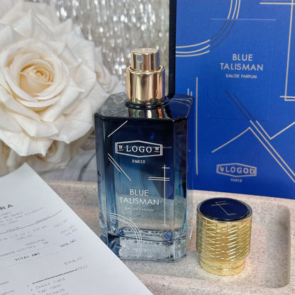 Vlogo Paris Blue Talisman Eau de Parfum bottle with a gold cap, displayed next to its blue and gold retail box and a small gold cylindrical container on a stone surface with a white rose in the background.