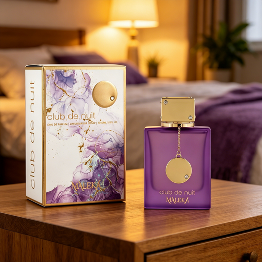 A purple rectangular perfume bottle with a gold cap stands beside its matching box on a wooden table in a softly lit bedroom. The packaging is white with purple floral artwork and gold text reading 'club de nuit' and 'MALEKA'.