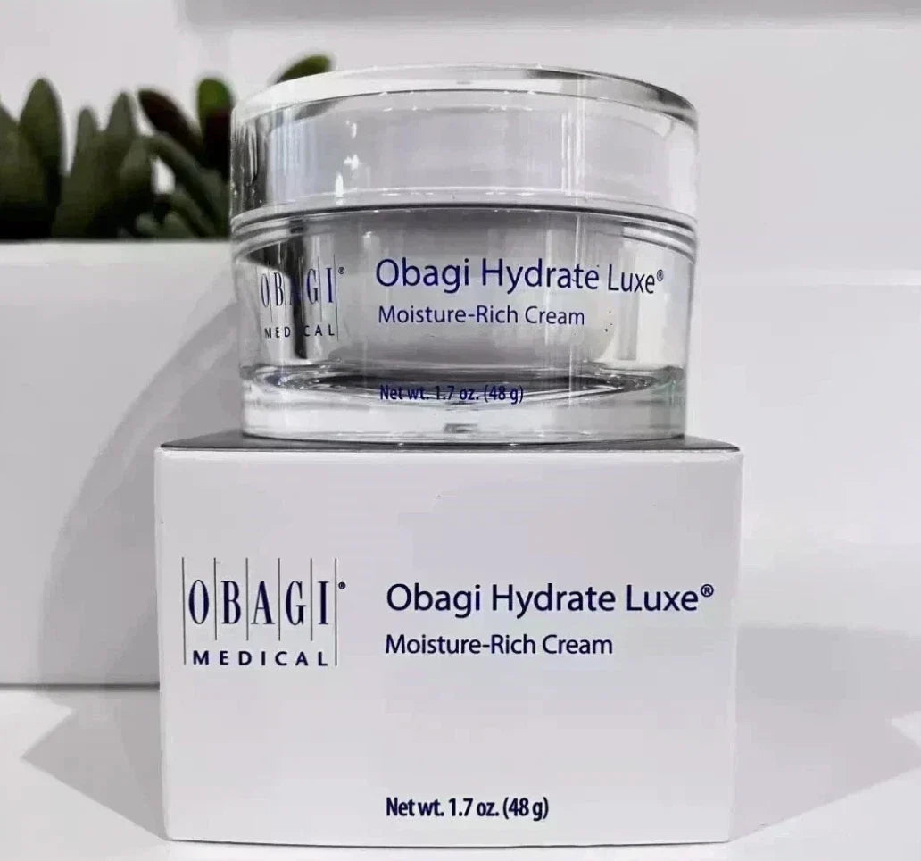 A clear cosmetic cream jar labeled Obagi Hydrate Luxe Moisture-Rich Cream, displayed on top of its white Obagi Medical box against a clean white background with a blurred green plant in the distance.