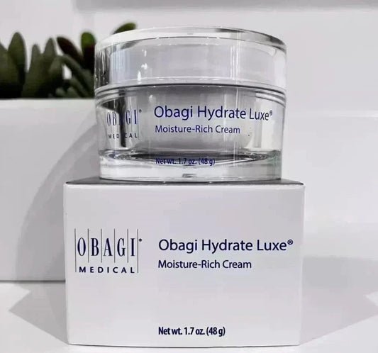 A clear cosmetic cream jar labeled Obagi Hydrate Luxe Moisture-Rich Cream, displayed on top of its white Obagi Medical box against a clean white background with a blurred green plant in the distance.