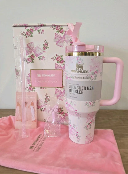 A pink floral Stanley Quencher H2.0 travel tumbler with a pink handle and lid, displayed alongside its branded gift box, a pink towel, and accessories on a wooden surface.