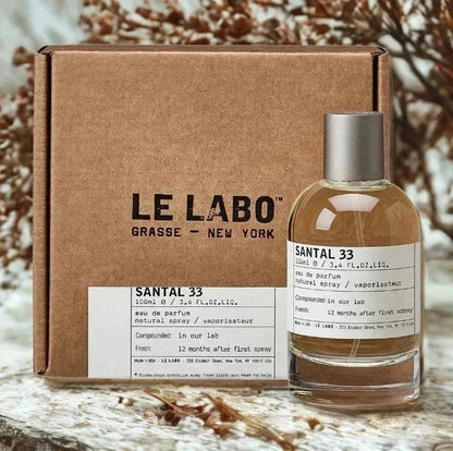 A bottle of Le Labo Santal 33 Eau de Parfum with a silver cap stands in front of its brown cardboard box. The bottle contains a warm amber liquid and has a white label with black text.