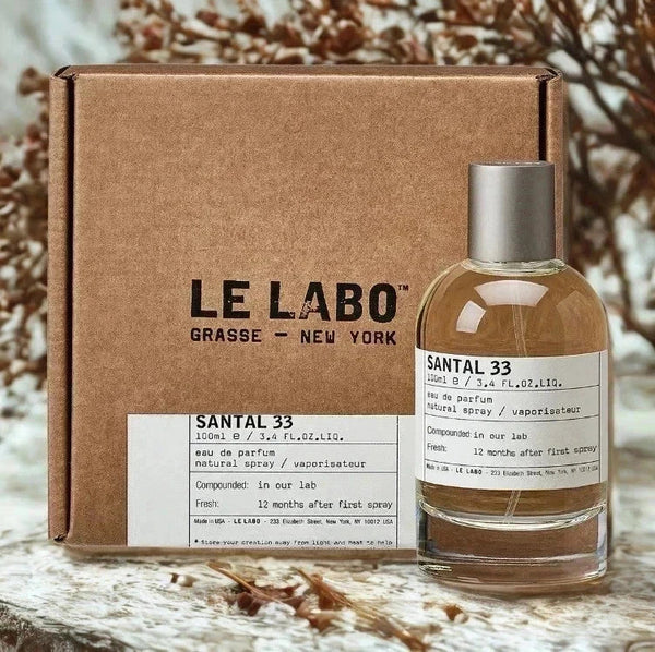 A bottle of Le Labo Santal 33 Eau de Parfum with a silver cap stands in front of its brown cardboard box. The bottle contains a warm amber liquid and has a white label with black text.
