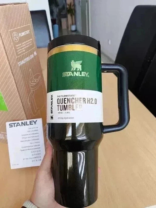 A hand holds a black Stanley Quencher H2.0 Tumbler with a green and white label, next to its brown packaging box and a small card on a wooden desk.