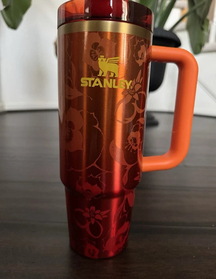 A red Stanley travel tumbler with a floral pattern, featuring a gold Stanley logo on the front and an orange handle, shown on a dark tabletop.